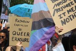 Image of a march at Amsterdam pride: figure holding up sign that reads: "Rainbow Capitalism = Queer Erasure"