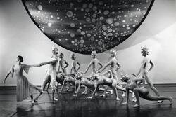 Twelve dancers onstage. Eleven wear leotards and hair caps with metallic balls attached to them, one wears a dress and leans away from the others.
