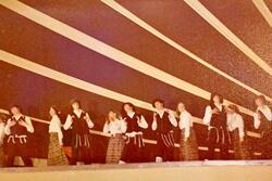 A group of dancers performing onstage, with the women wearing long dresses and headscarfs, and the men wearing black vests, hats, and trousers, white shirts, and tallit katan