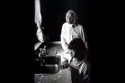 Portrait of an older woman with a white headscarf and a young girl, standing by a kitchen sink
