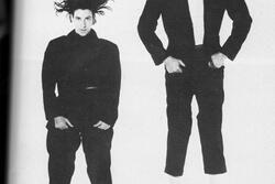 Two dancers in black pants, jackets, and shoes, jumping in the air at different heights