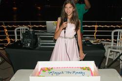 Maddy Pollack speaking at her Bat Mitzvah party, microphone in hand. Cake on a table in front of her with text "Mazel Tov, Maddy."