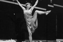 Ruth Eshel standing in a pile of loose wool, with one leg and both arms lifted high, wearing a thick iron circlet around her head.