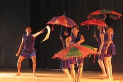 A group of dancers on stage, all wearing loose blue/purple velvet shorts and shirts. Four are holding colorful red, red/purple, and red/green umbrellas, one holds a horse hair whip