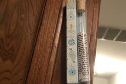 Mezuzah mounted on wooden door frame. Scroll visible, in a test tube-like vial corked on light blue wood decorated with pomegranates and Star of Davids.