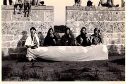 Four dancers sitting at a white cloth stretched between them in front of stone walls. Children sit on top of the walls.