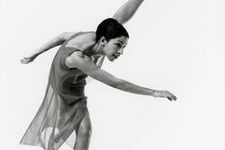 Ze'eva Cohen, dressed in a skirted leotard, striking a dance post with one foot on the ground and the other lifted above her head
