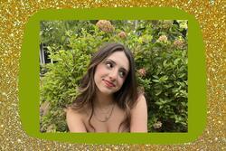 Image of Eva smiling and wearing gold necklaces with flowers in background