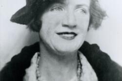 black and white photograph of a woman wearing a hat and a dark jacket and a necklance, looking off to the side with a slight smile on her face