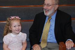 Six year old in pink dress seated next to elderly man dressed in a suit, sitting in pews at a synagogue