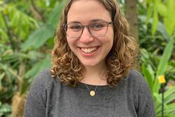 Young woman with brown curly hair and glasses wearing dark gray shirt and posing in front of trees