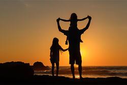 Silhouette of children with adult in front of beach sunset, one of the children is on the adult's shoulders and the other is next to them.