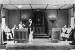 A picture of a bimah, with two men and two women sitting on either side, and a man in a kippah and prayer shawl standing by a podium, and a young woman with flowers pinned to her dress speaking at the podium