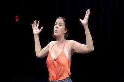 Woman in a theatrical performance, wearing orange tank stop and with arms in "hands up" position