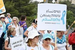 Two women wearing white and marching  holding a sign that says "Women Wage Peace" in English, Hebrew, and Arabic