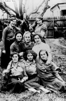 Eight women in casual blouses and skirts, posing in front of a tree