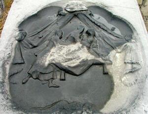 A gray stone, carved with the image of three women, a bed, and parted curtains