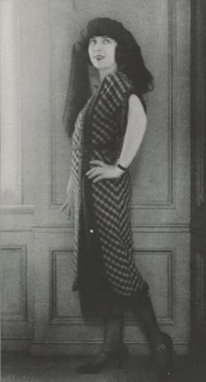 Sally Milgrim posing in a calf-length dress with a plaid pattern, black mary jane shoes, and a turban-style hat with fabric draped from it
