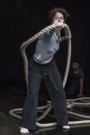 Tatiana Zugazagoitia, standing on stage, wearing loose black pants and a grey shirt, holding a rope, with another dancer kneeling on the stage behind her