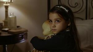 A young girl sitting on a bed looking to camera over her shoulder while holding a doll