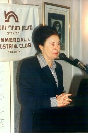 Ora Namir standing in front of a microphone with a sign behind her with Hebrew and English