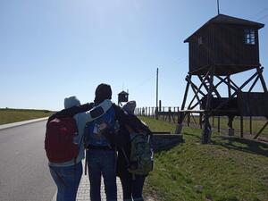 Three figures, arms over shoulders, walking on a paved road. Guard towers lined alongside them.