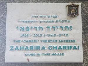 A white plaque with blue lettering in Hebrew and English. The English reads "1929-2013. The 'Cameri' Theater Actress Zaharira Charifai lived in this house."