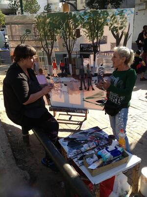 Zoya Cherkassky seated outdoors in front of a large canvas, talking to another woman