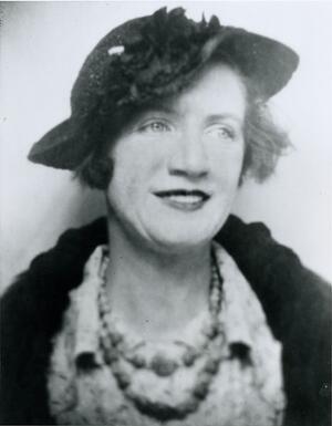 black and white photograph of a woman wearing a hat and a dark jacket and a necklance, looking off to the side with a slight smile on her face