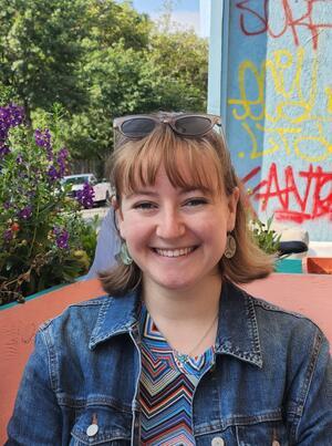 Photo of young women with chin length brown hair and bangs, wearing denim jacket and sunglasses on head