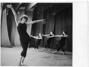 Four dancers wearing black tank tops and footless leggings. One is posing with her arms in the air. The other three, further behind her and to the right, have their legs crossed and are leaning back.