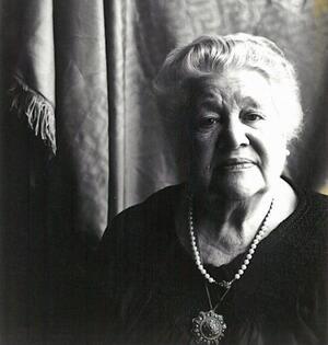 Black and white phtoograph of a woman with white hair wearing a dark dress looking pensively at the camera
