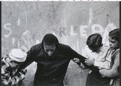 Black boy standing in front of wall with head bowed, arms being held by several white boys