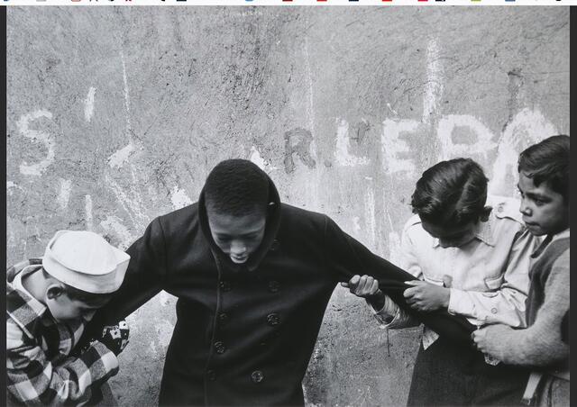 Black boy standing in front of wall with head bowed, arms being held by several white boys