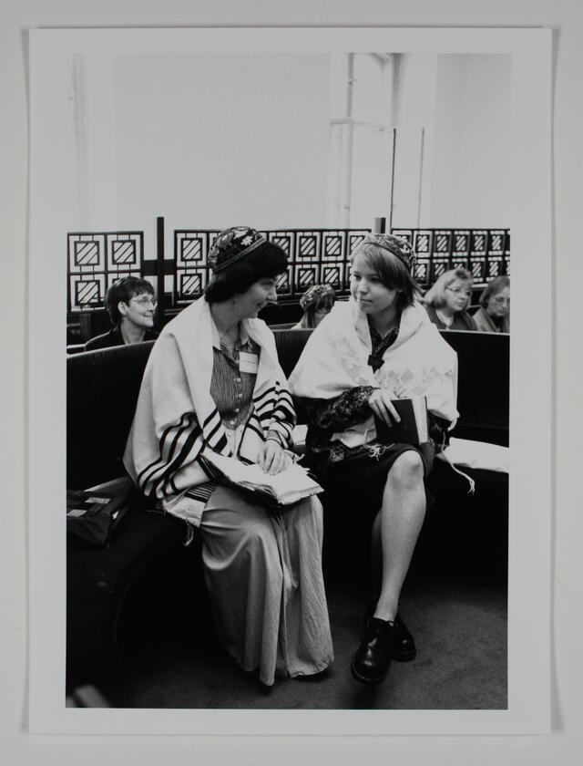 Two women sitting on a bench, wearing prayer shawls and kippot and holding prayer books