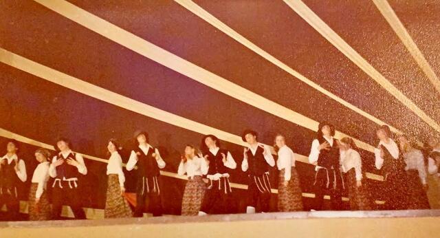 A group of dancers performing onstage, with the women wearing long dresses and headscarfs, and the men wearing black vests, hats, and trousers, white shirts, and tallit katan