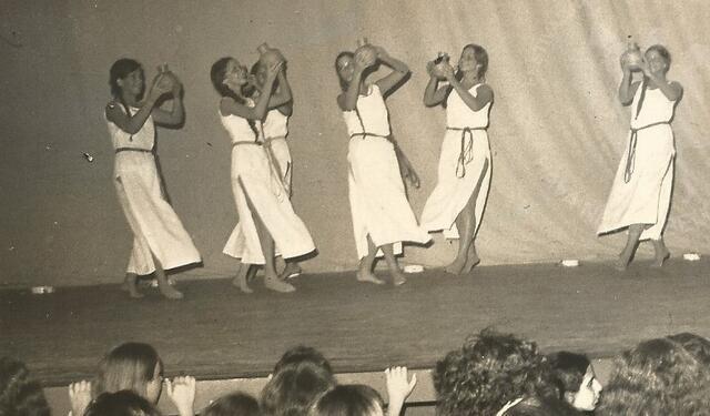 A group of six women, barefoot, wearing long white tunics, and holding up clay pots as they dance onstage