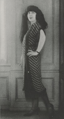 Sally Milgrim posing in a calf-length dress with a plaid pattern, black mary jane shoes, and a turban-style hat with fabric draped from it