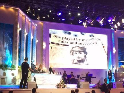 Over a stage with a panel of people and musicians an image of a newspaper with the headline She played by men-made rules and succeeded is projected
