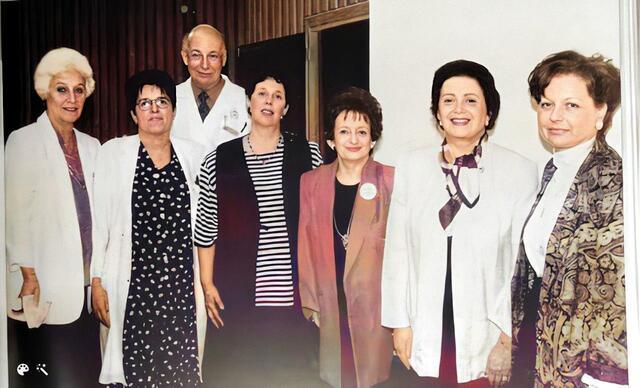 Six women and one man standing together, four wearing white doctor's coats