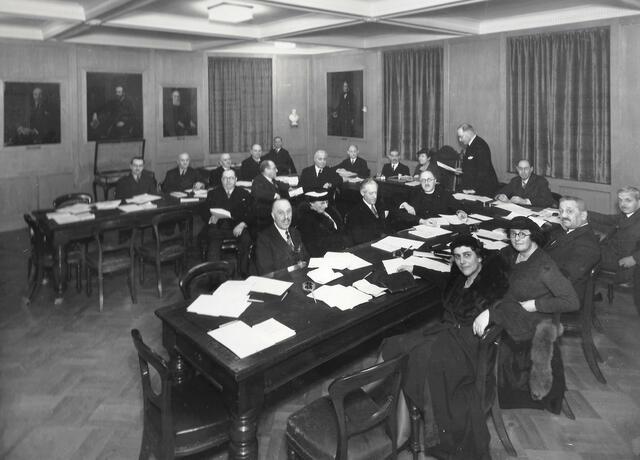 A group of men in suits and four women sitting at three rectangular tables