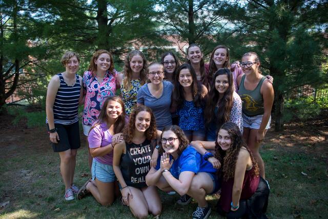 14 young women smiling for a group photo with some standing and some squatting, outdoors surrounded by trees