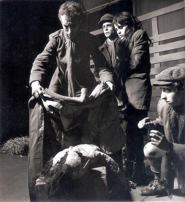 Four actors in tattered clothing. One man drapes a coat over a dummy while the others watch