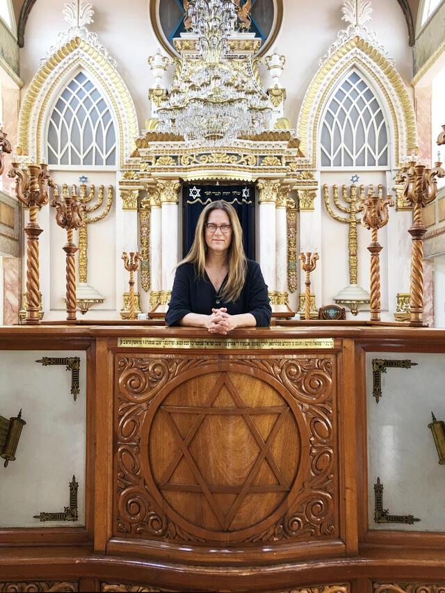 Mónica Unikel-Fasja standing before the Torah ark in an elaborately decorated synagogue with carved candelabras, menorahs, and columns.