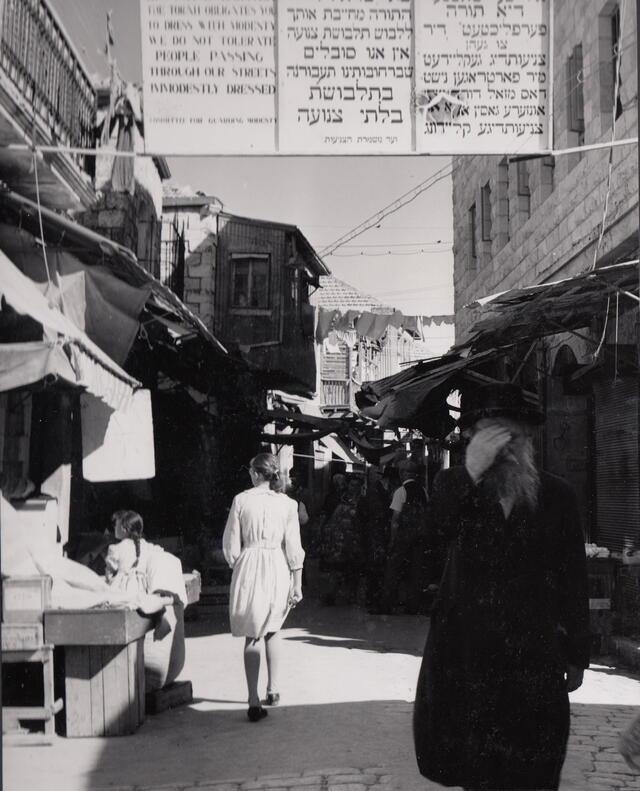 Photo of woman wearing a short dress in Mea Shearim, Israel, underneath a sign mandating modest dress. A man covers his eyes as she passes.