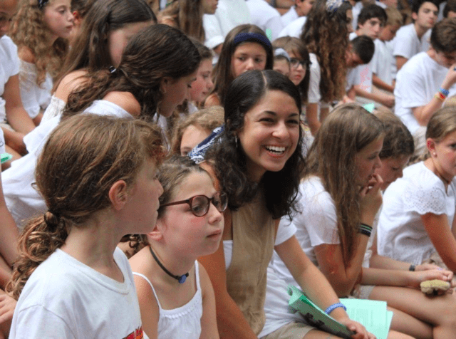 Camp counselor surrounded by campers, all wearing white. Sitting on benches during services.