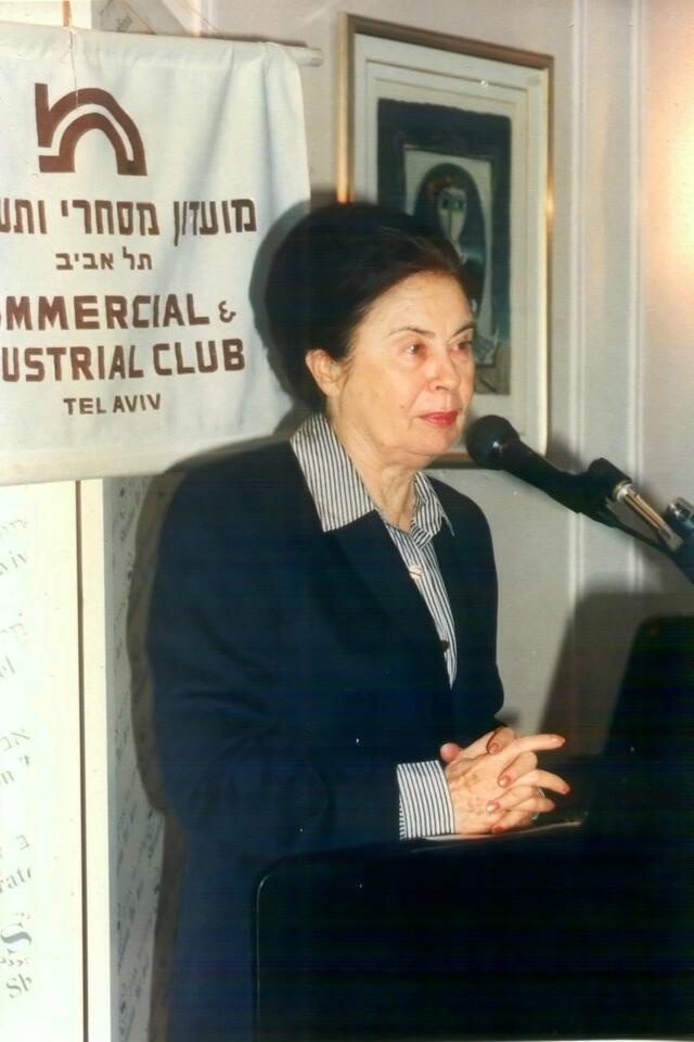 Ora Namir standing in front of a microphone with a sign behind her with Hebrew and English