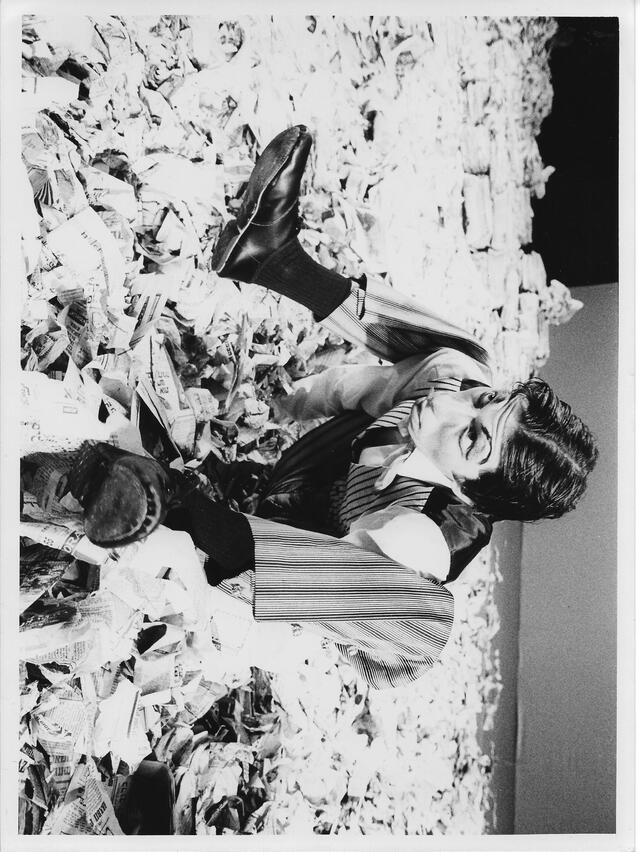 Dancer in costume and make up for Headlines posed with newspapers on ground 1979
