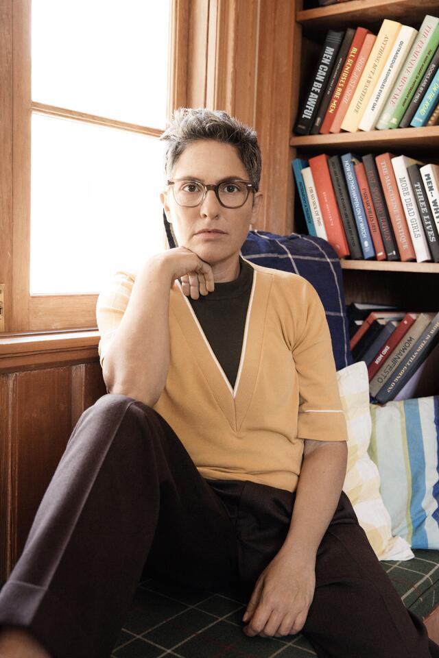 Joey Soloway wearing dark pants, yellow top, and glasses, seated in front of a window and a bookcase with right arm propped up on bent right leg