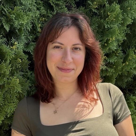 Emma Breitman stands smiling in front of green foliage. She has reddish brown hair and is wearing an olive green shirt.